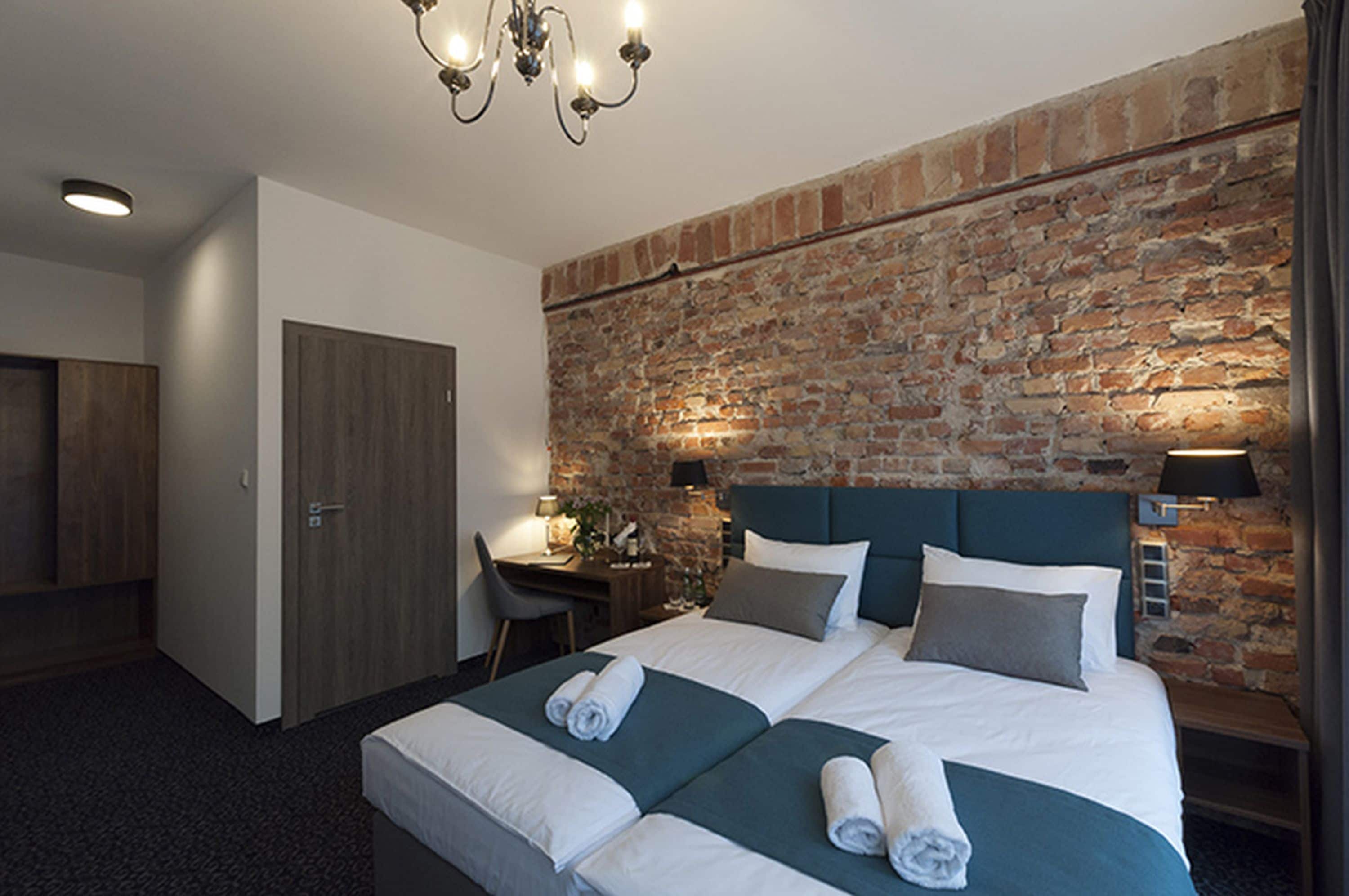 A hotel room with a brick wall, a large bed with white bedding, a desk with a chair, and a chandelier.