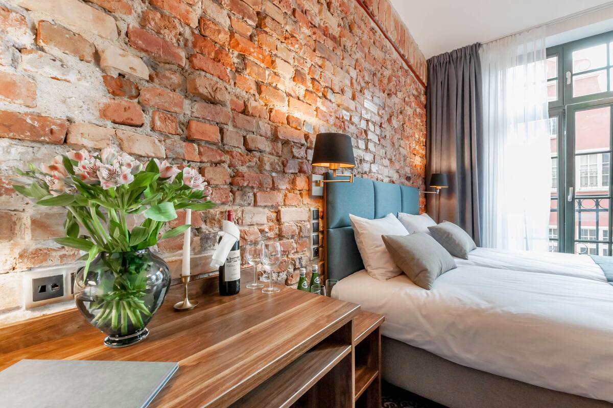A hotel room with a brick wall, a bed with white bedding, a bedside table with a vase of flowers, and a lamp.