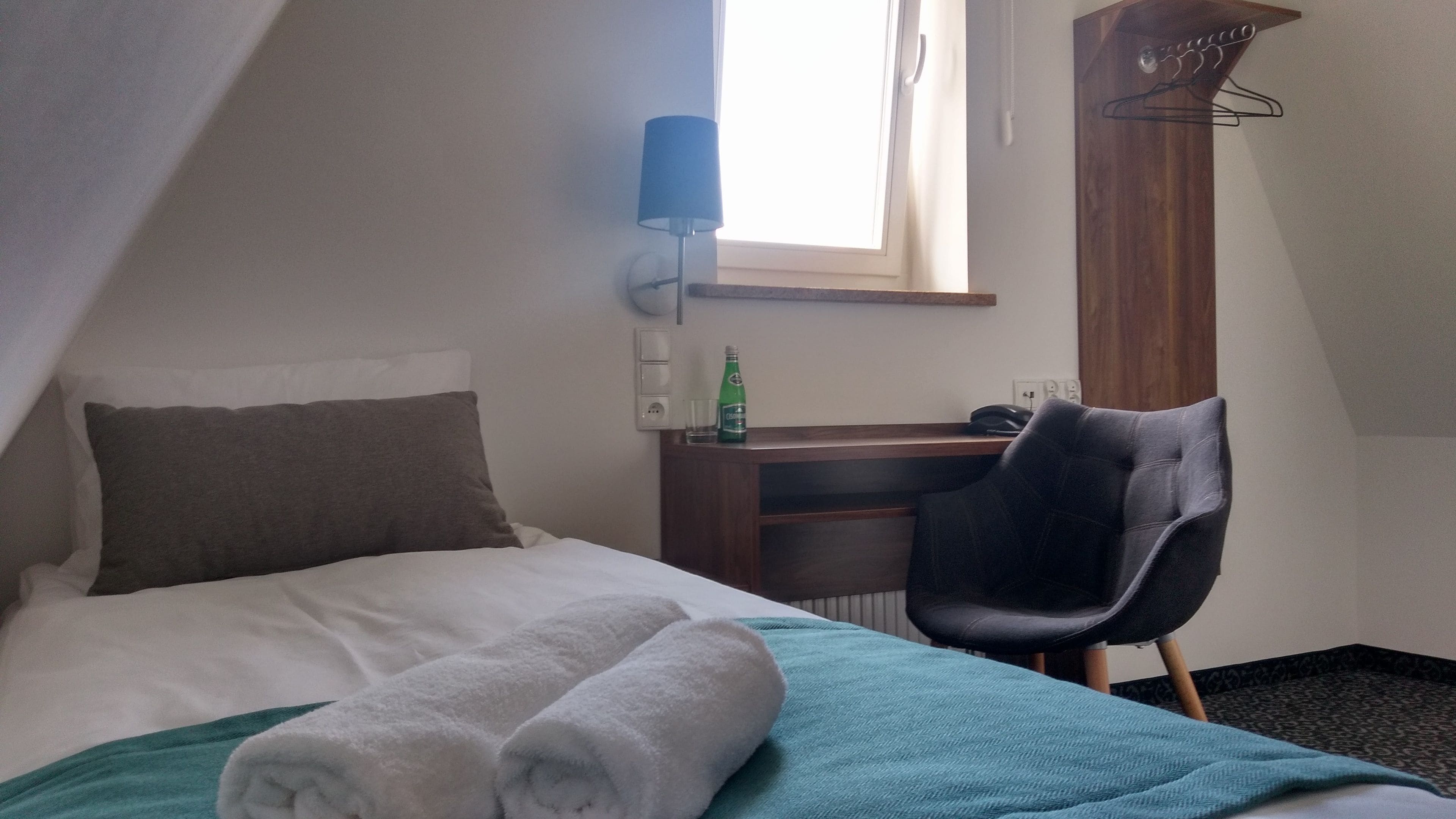 A hotel room with a bed, a desk, a chair, a lamp, and a window.