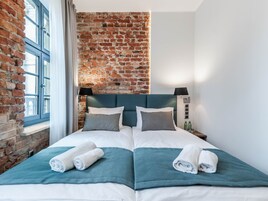 A double bed with teal bedding, two pillows, and rolled towels. A brick wall and a window with curtains are visible.