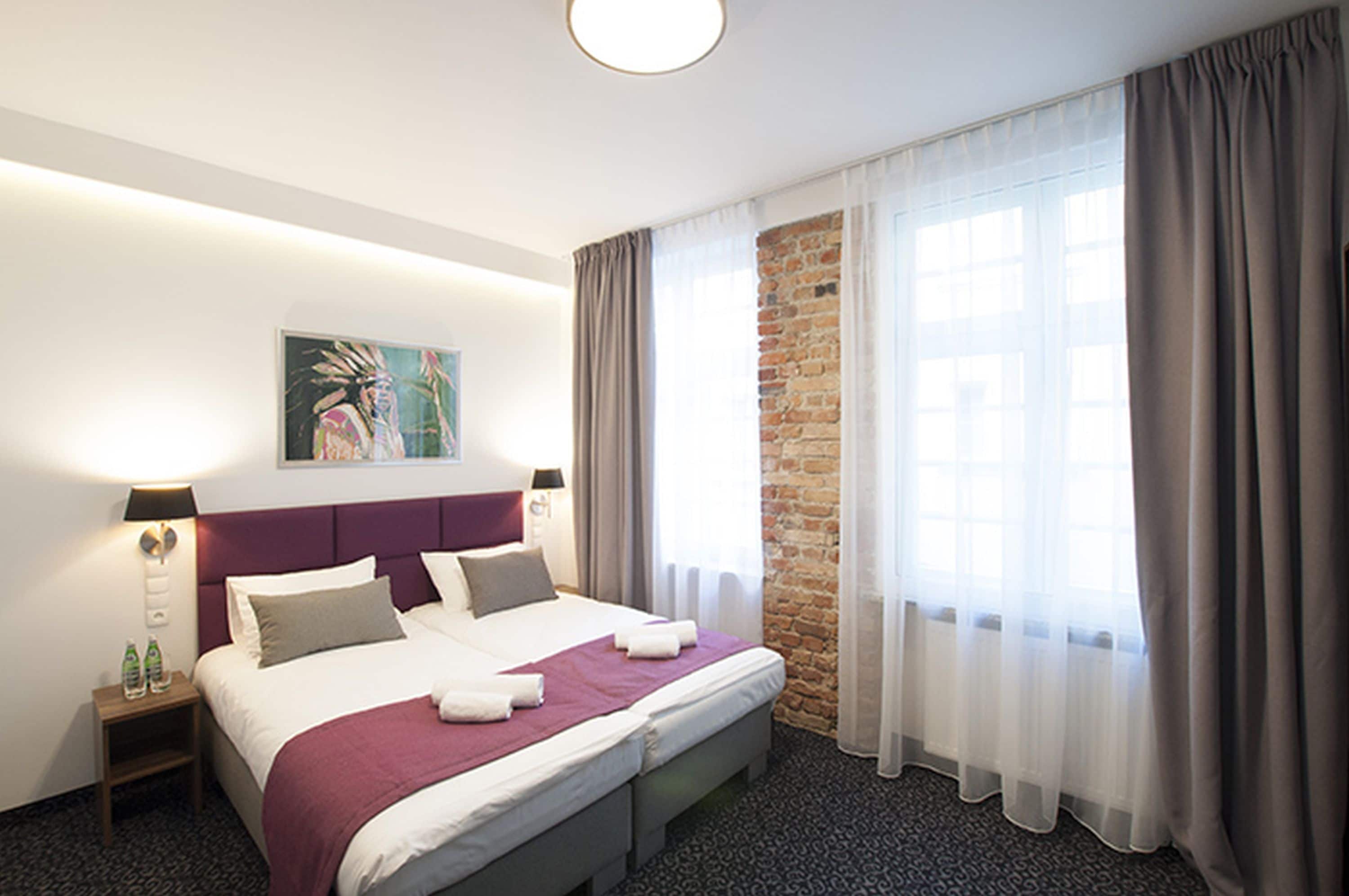 Una habitación de hotel con una cama grande, cabecera, mesita de noche, un cuadro, pared de ladrillo y una ventana con cortinas.