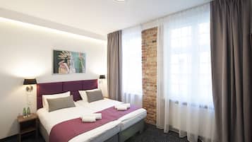 Una habitación de hotel con una cama grande, cabecera, mesita de noche, un cuadro, pared de ladrillo y una ventana con cortinas.