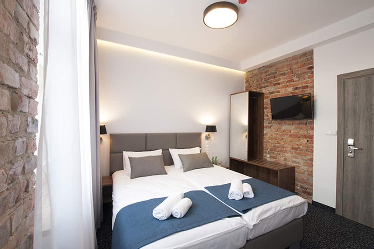 A modern hotel room with a brick wall, a large bed with two pillows, a flat-screen TV mounted on the wall, and a wooden door.