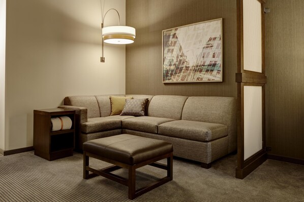 A corner living room with a sectional sofa, a small wooden table, a bench, a wall-mounted floor lamp, and a framed picture.