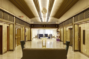 Een ruime lobby met een centrale zithoek, een klok aan het plafond en decoratieve wandpanelen.