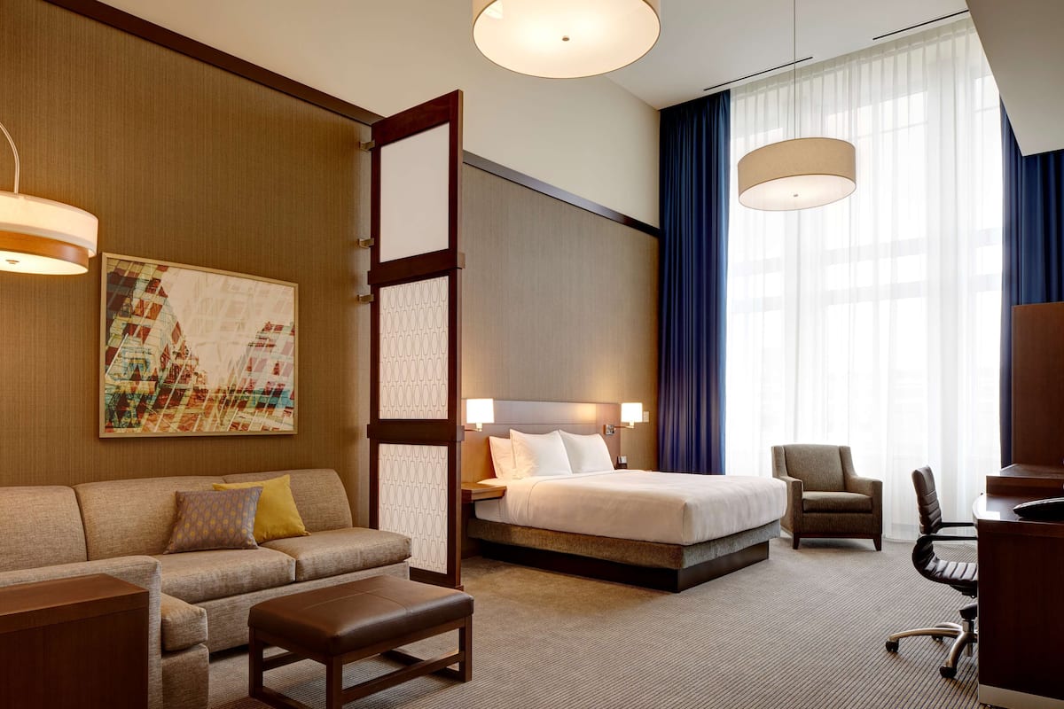 Hotelkamer met een bed, een bank, een salontafel, een bureau en een stoel. Twee hanglampen en een groot raam met gordijnen.