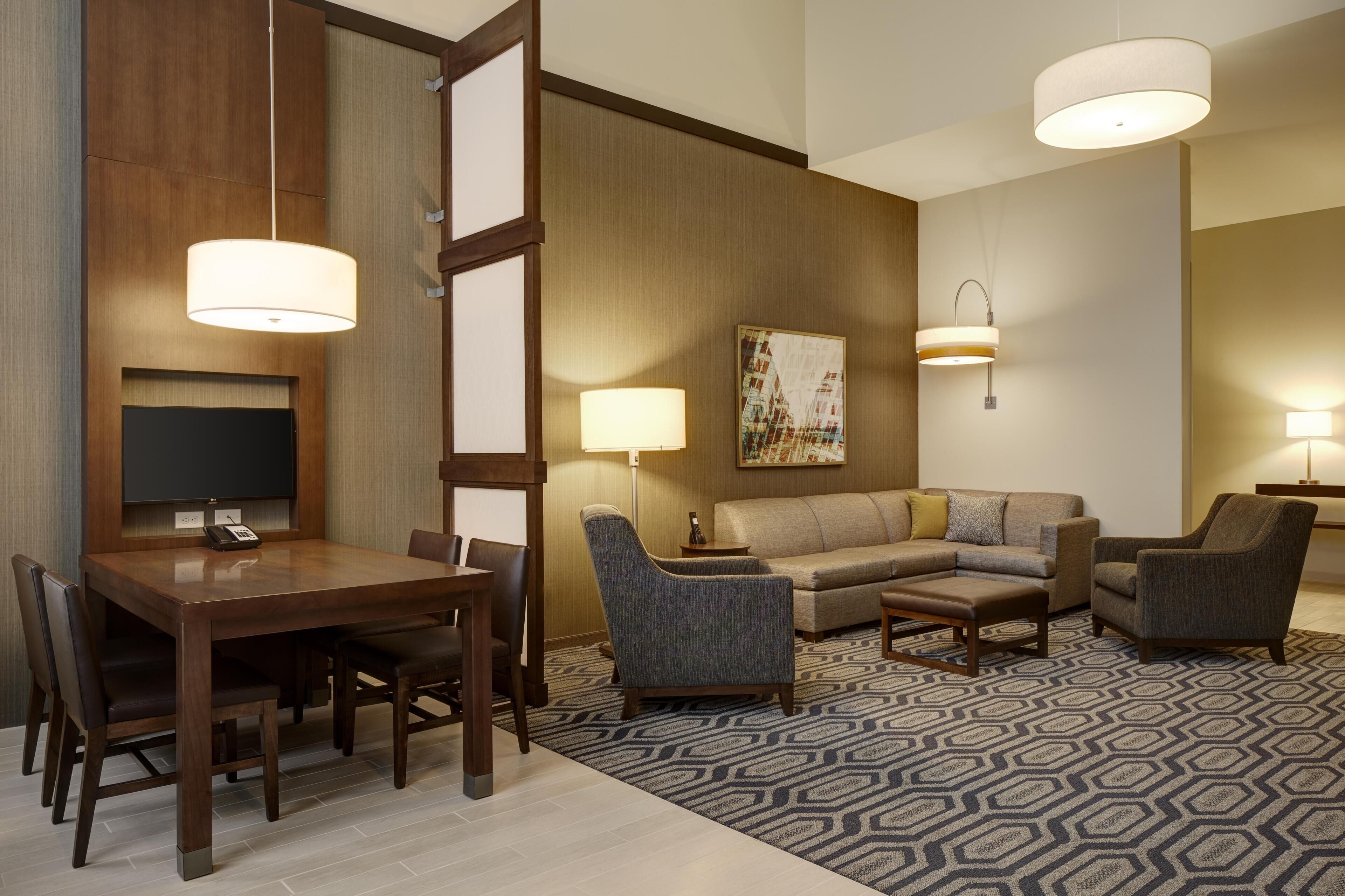 A hotel room with a dining table, chairs, a sofa, and a patterned rug.