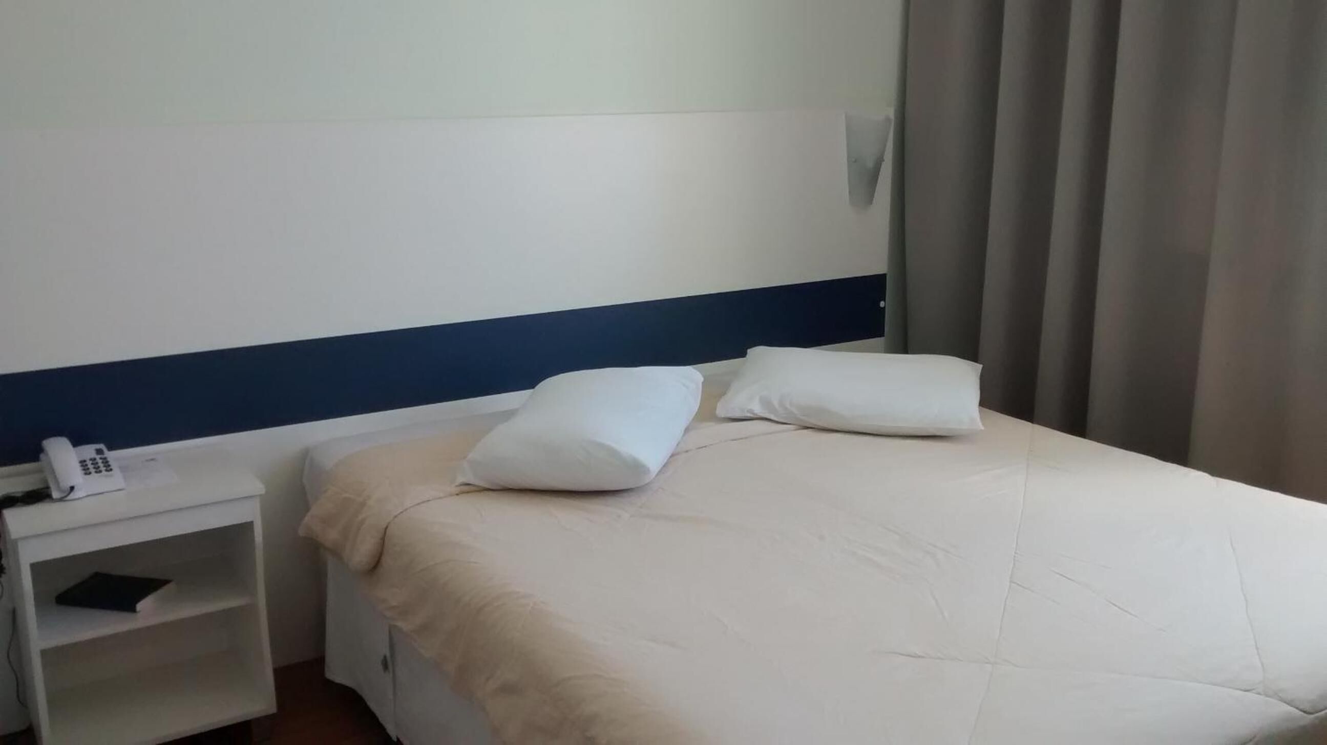 A hotel room with a bed, two pillows, a bedside table with a phone, and a shelf with a book.