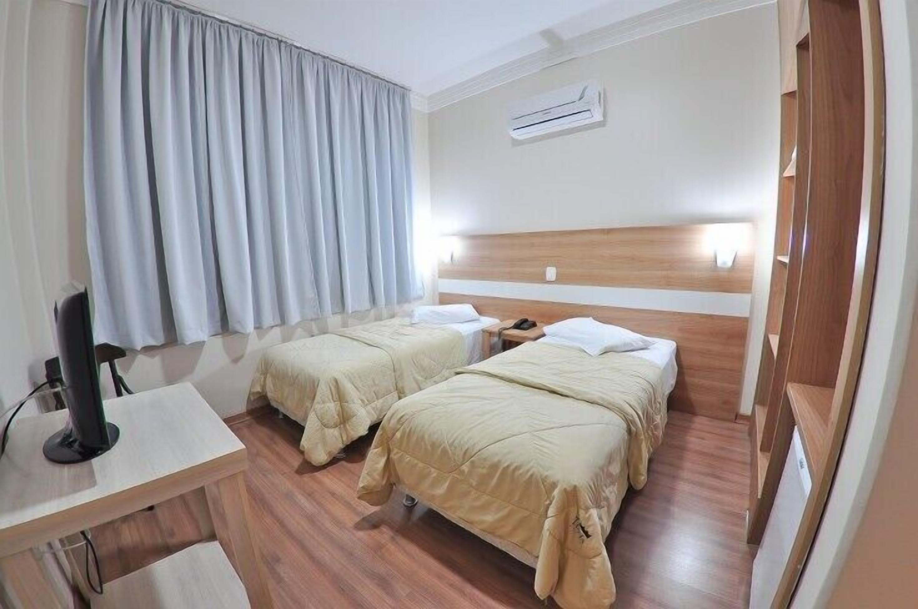 Habitación de hotel con dos camas, un escritorio con computadora, aire acondicionado y una ventana con cortinas.