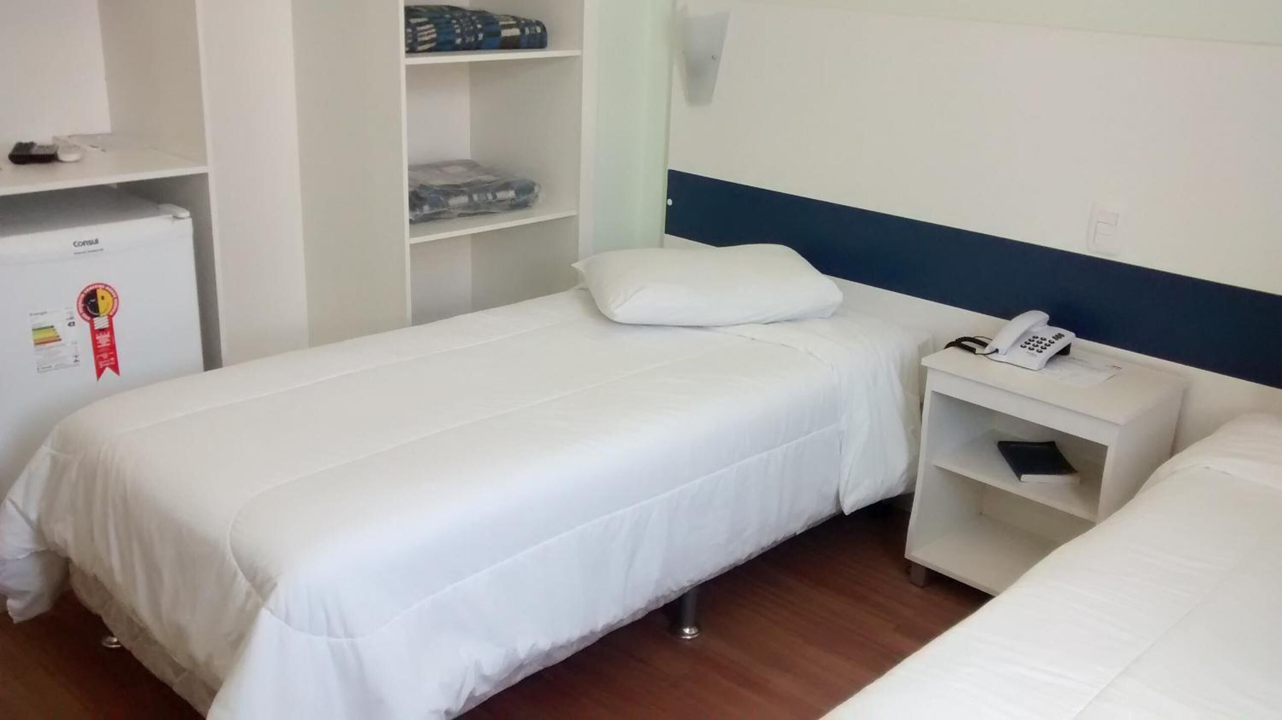 Ein Zimmer mit einem Einzelbett, einem kleinen Nachttisch mit Telefon, einem weißen Kleiderschrank und einem Kühlschrank.