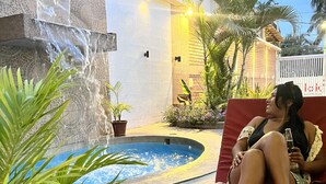 A person relaxing on a red lounge chair by a pool with a waterfall feature, surrounded by potted plants and palm trees. - Hotel Aryia (Máncora)