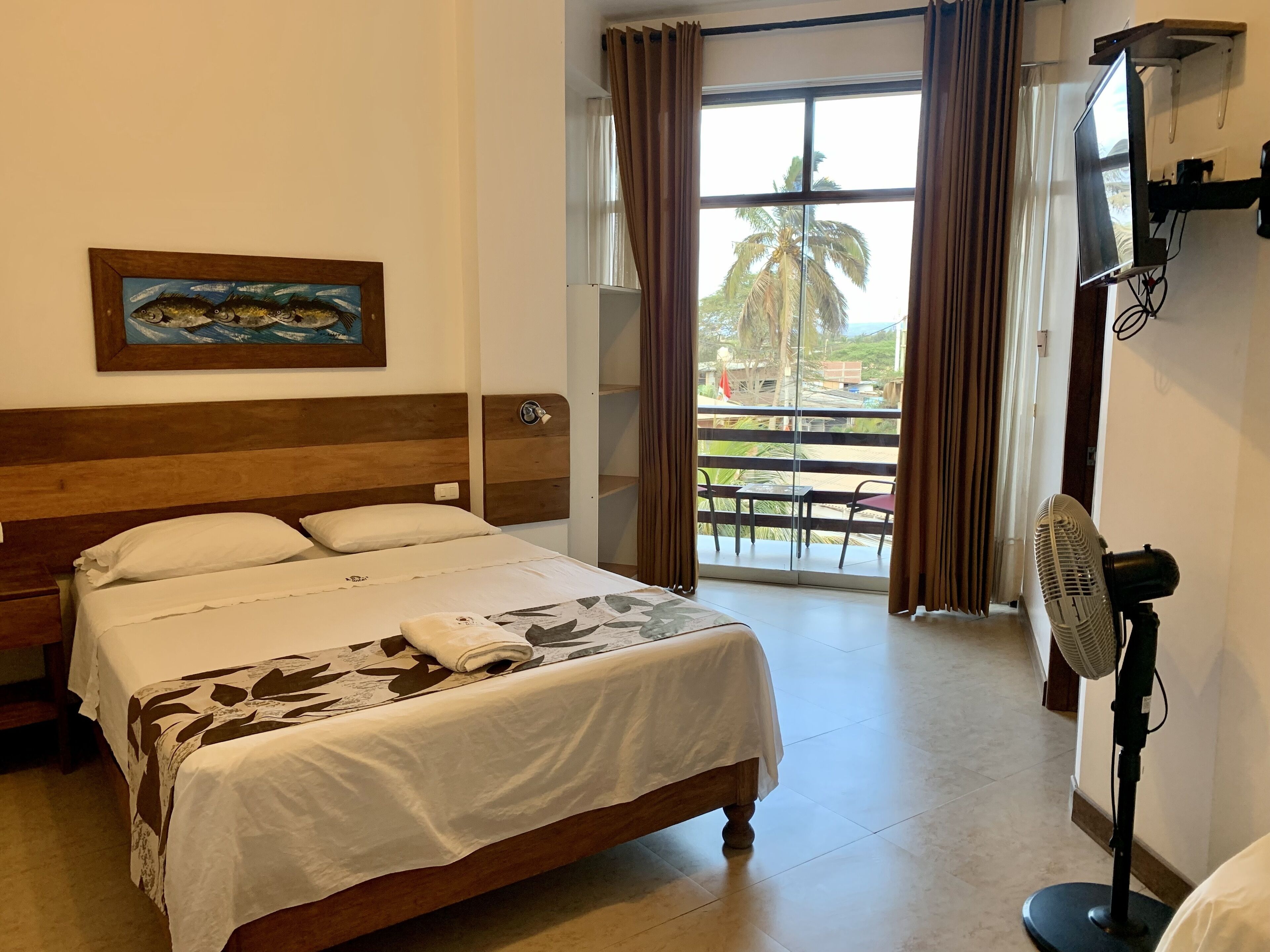 Habitación de hotel con cama, un cuadro, televisor y balcón con vista a palmeras.