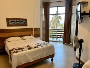 A hotel room with a bed, a painting, a TV, and a balcony with a view of palm trees. - Hotel Aryia (Máncora)