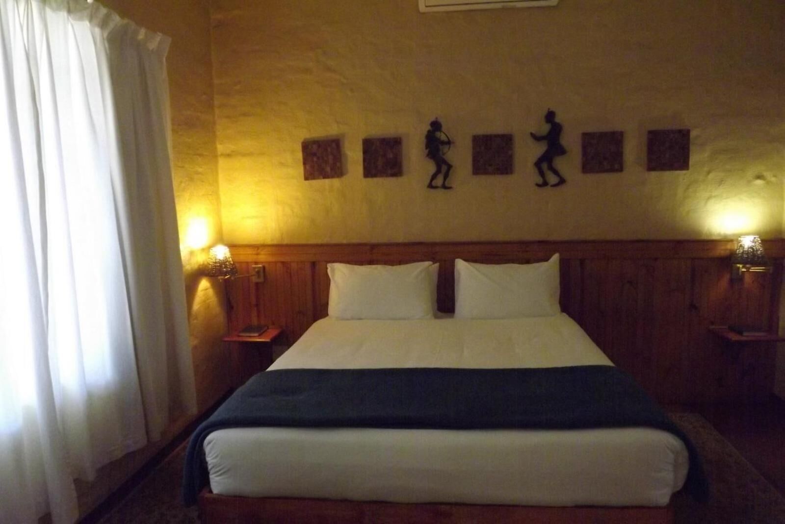 Habitación de hotel con una cama grande, dos mesitas de noche con lámparas, un cabecero de madera y una pared con tres siluetas enmarcadas.