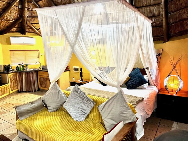 A room with a bed under a canopy, a bedside table, a lamp, and a view of the kitchen. - Manzini Swazi King Chalets (Nkomazi)