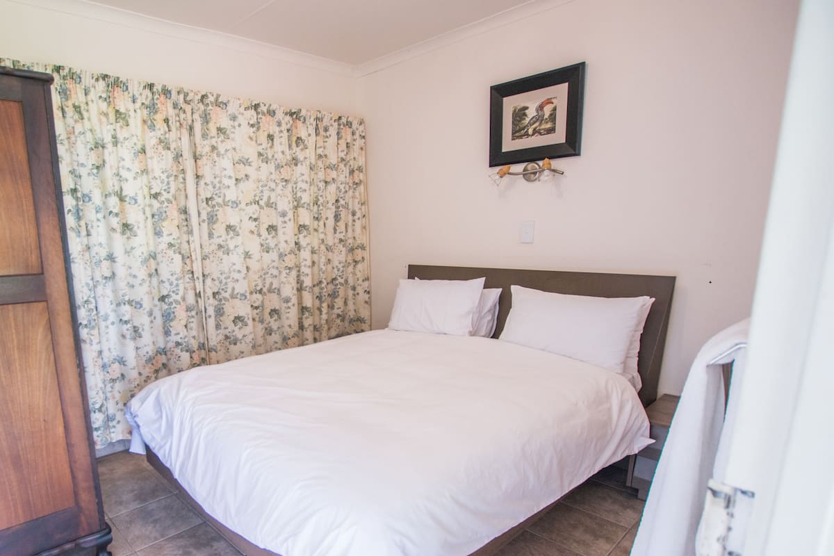 A neatly made bed with white linens in a room with a wooden headboard, floral curtains, and a framed picture on the wall.