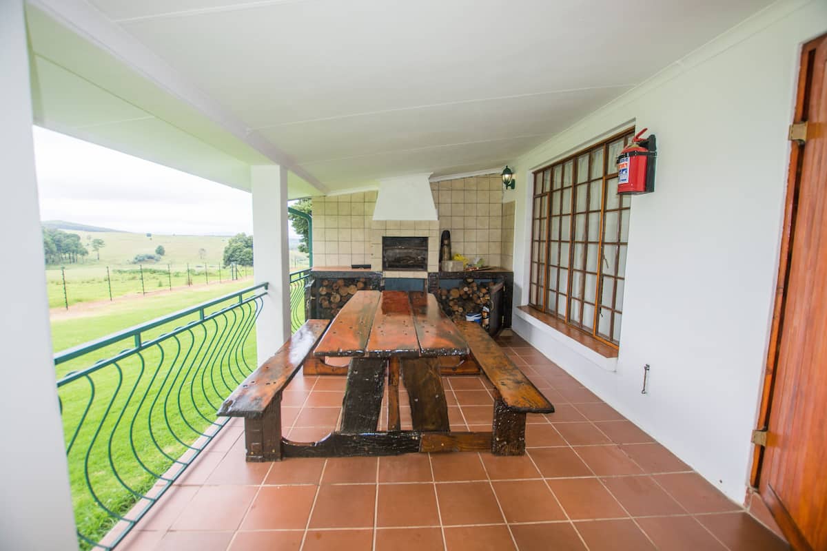 A covered patio with a wooden table and benches, a tiled wall with a fireplace, and a view of a green landscape.