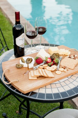 A picnic setup with a bottle of wine, glasses, cheese, crackers, and fruits by a poolside table. - Four Palms Accommodation (Cape Town)