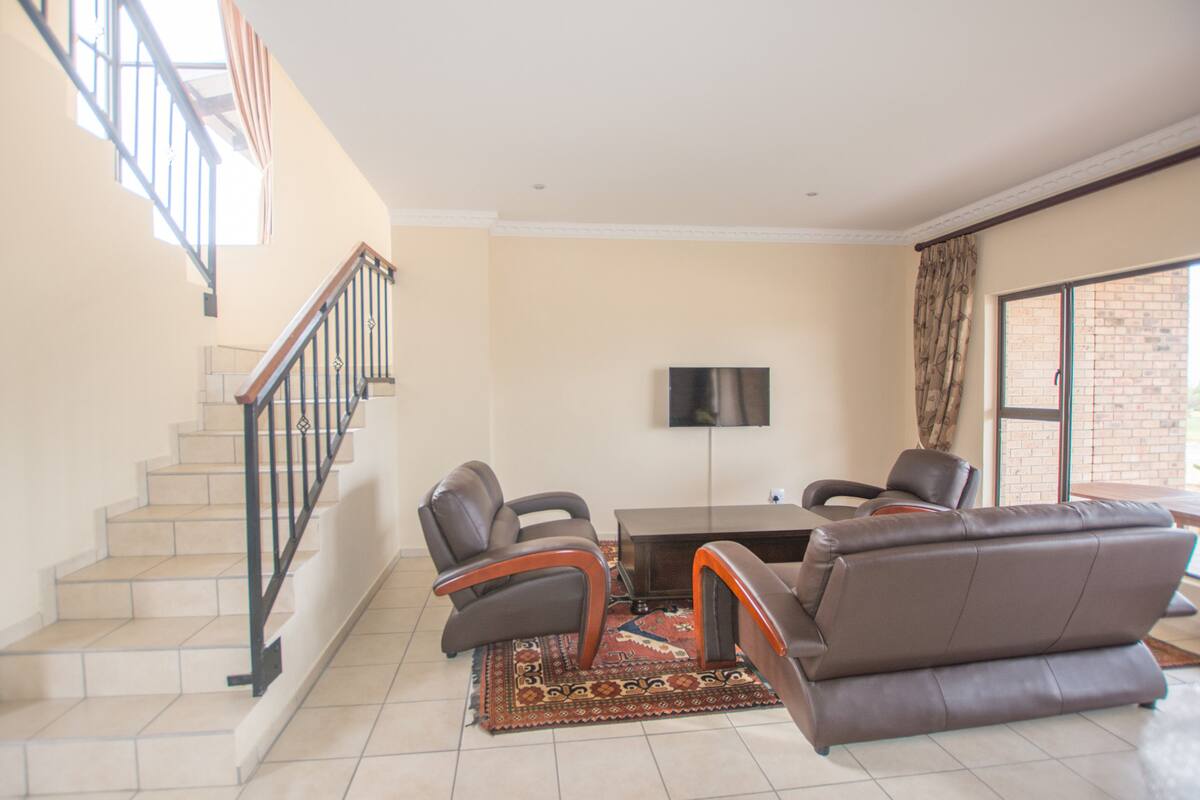 A living room with a sofa, two armchairs, a coffee table, and a TV mounted on the wall. There is a staircase with a railing leading to an upper floor.