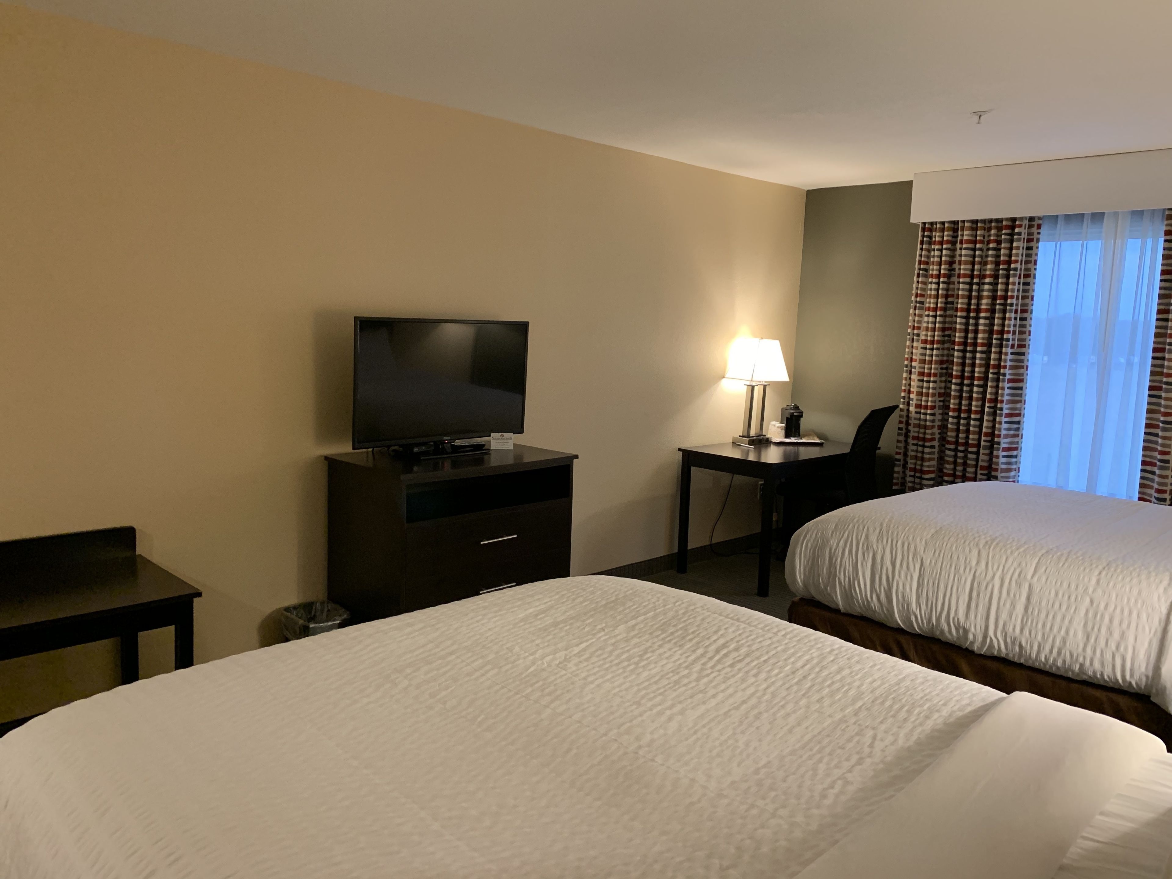 A hotel room with two beds, a TV on a dresser, a desk with a lamp, and a window with curtains.