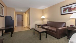 A hotel room with a brown leather sofa, a small table, a lamp, and a bed. - Days Inn & Suites by Wyndham Florence/Jackson Area (Florence)