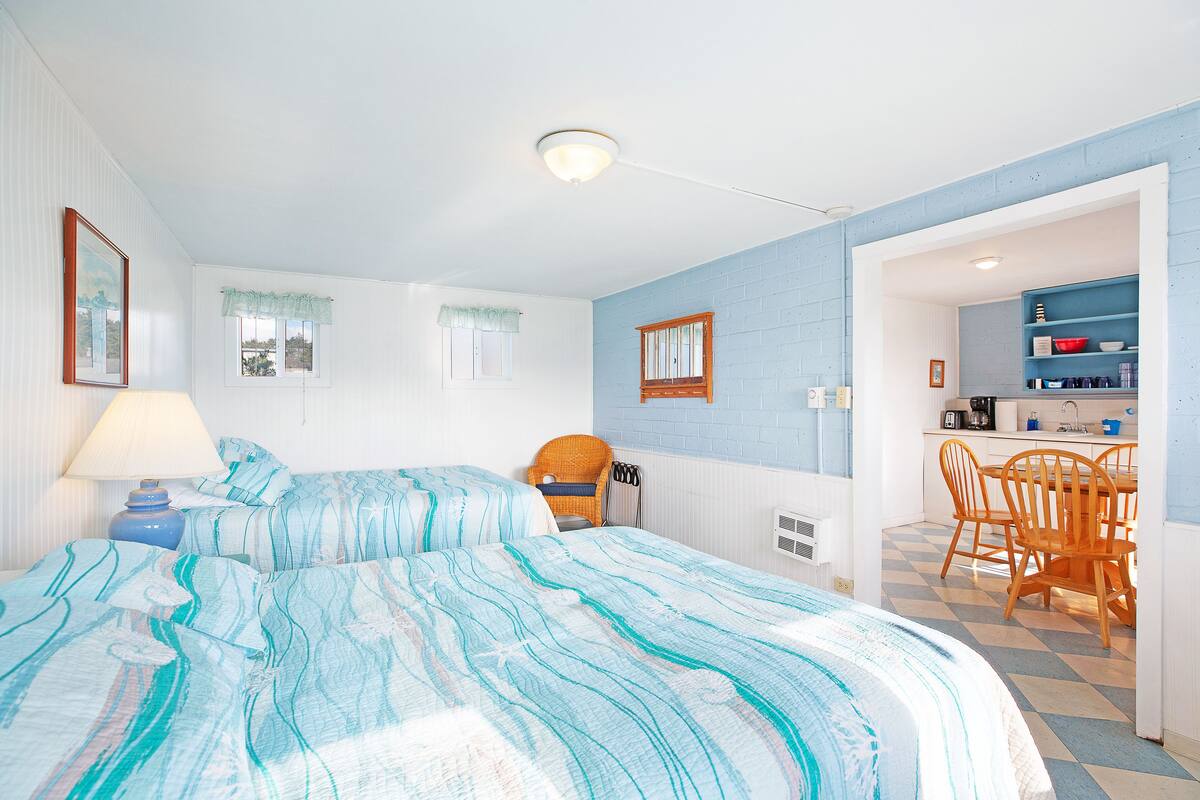 A bedroom with two beds, a chair, a dining table, and a kitchenette.