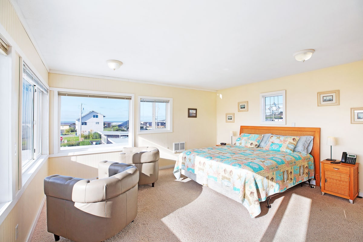 A bedroom with a large bed, two armchairs, a nightstand, and a window with a view of houses.