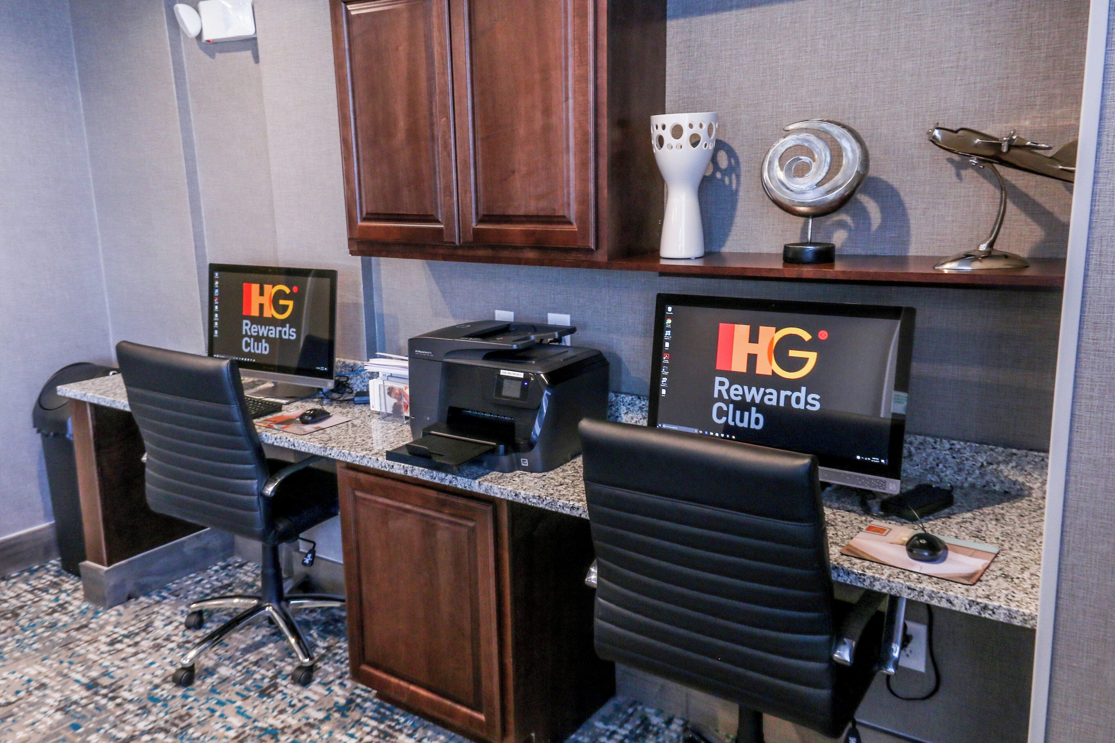 Habitación de hotel con escritorio, dos sillas, una impresora, un monitor de computadora y un letrero que indica el HG Rewards Club.