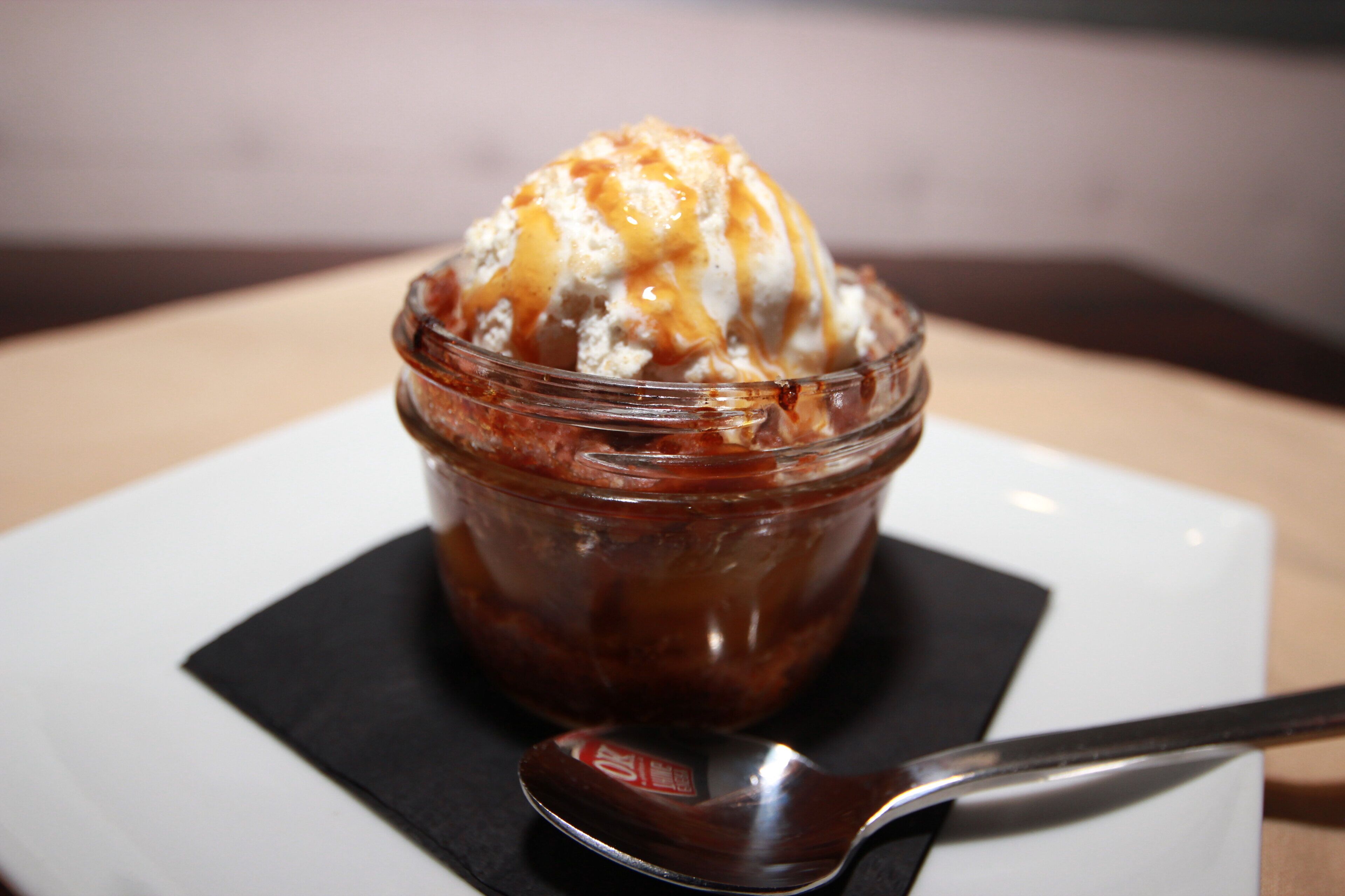 A dessert in a glass jar with caramel sauce on a white plate.