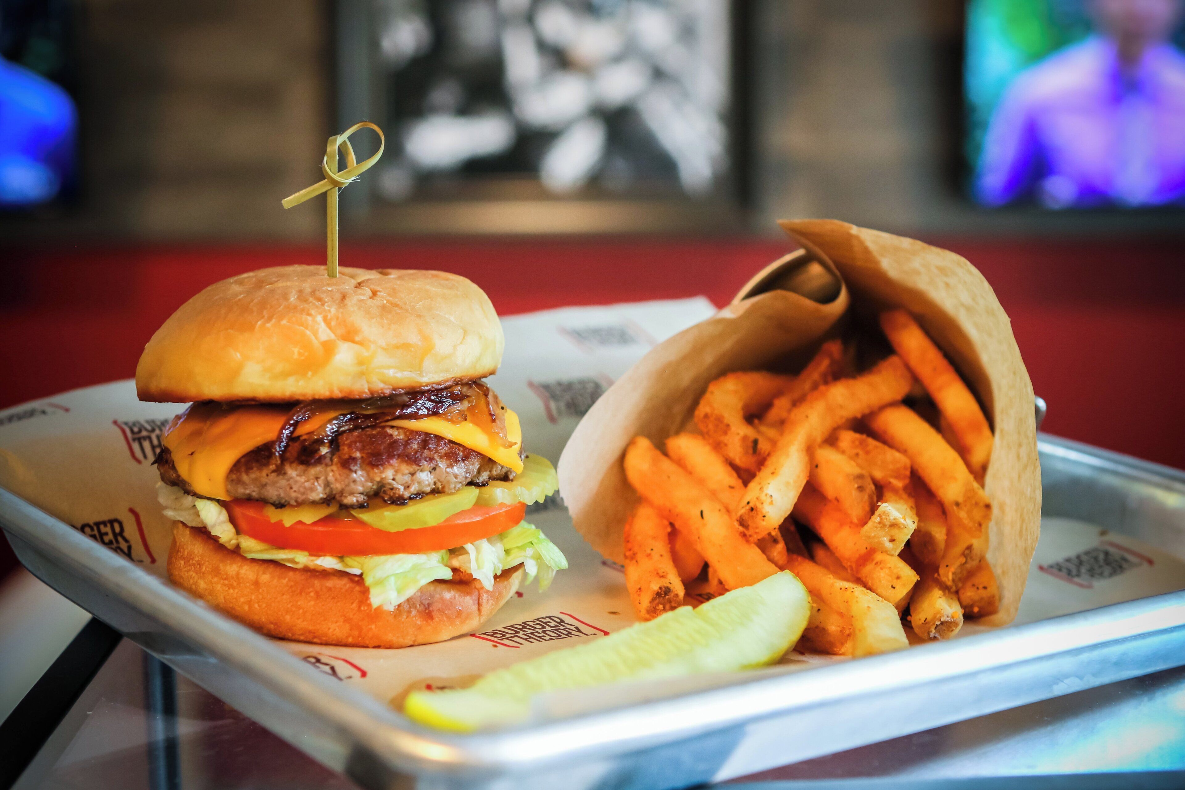 A burger with a toothpick on top, a side of fries, and a pickle on a tray.