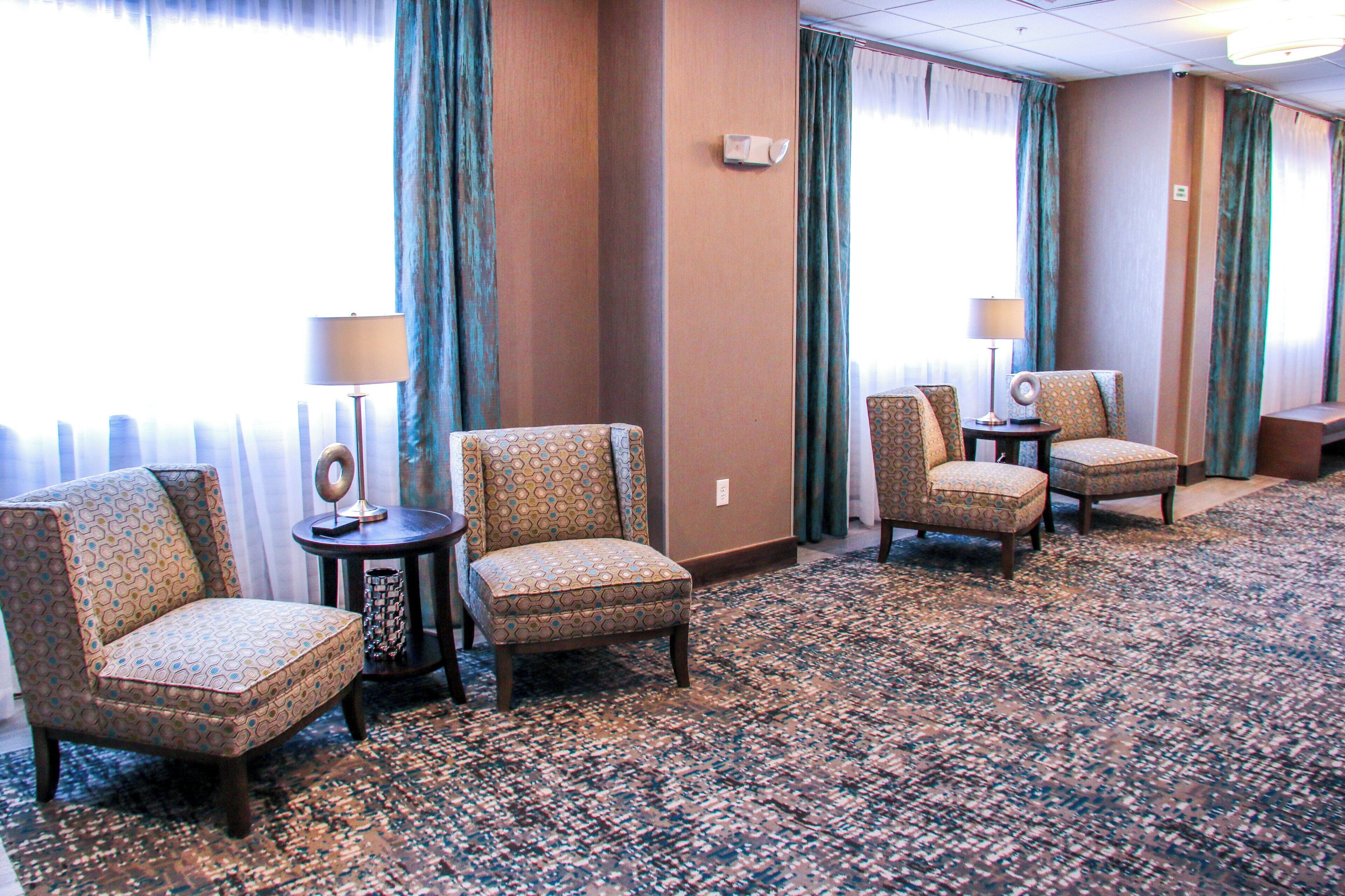 A hotel room with a carpeted floor, two armchairs, a small table with a lamp, and a view of the outdoors through a window with curtains.