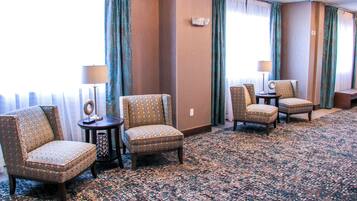 A hotel room with a carpeted floor, two armchairs, a small table with a lamp, and a view of the outdoors through a window with curtains.