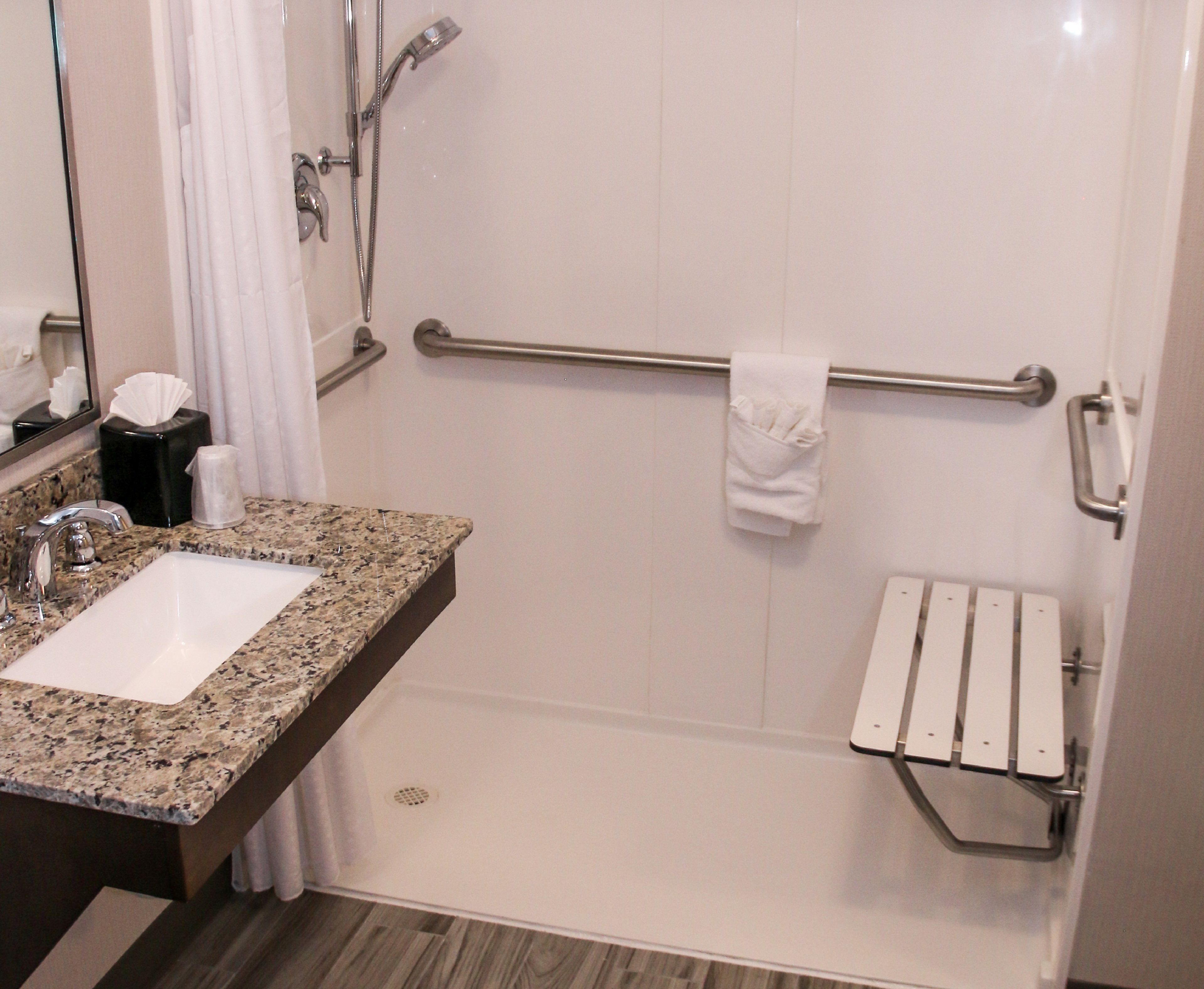 A bathroom with a granite countertop sink, a mirror, a towel rack, a shower with a grab bar, and a toilet paper holder.