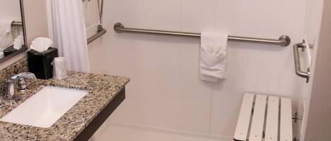 A bathroom with a granite countertop sink, a mirror, a towel rack, a shower with a grab bar, and a toilet paper holder.