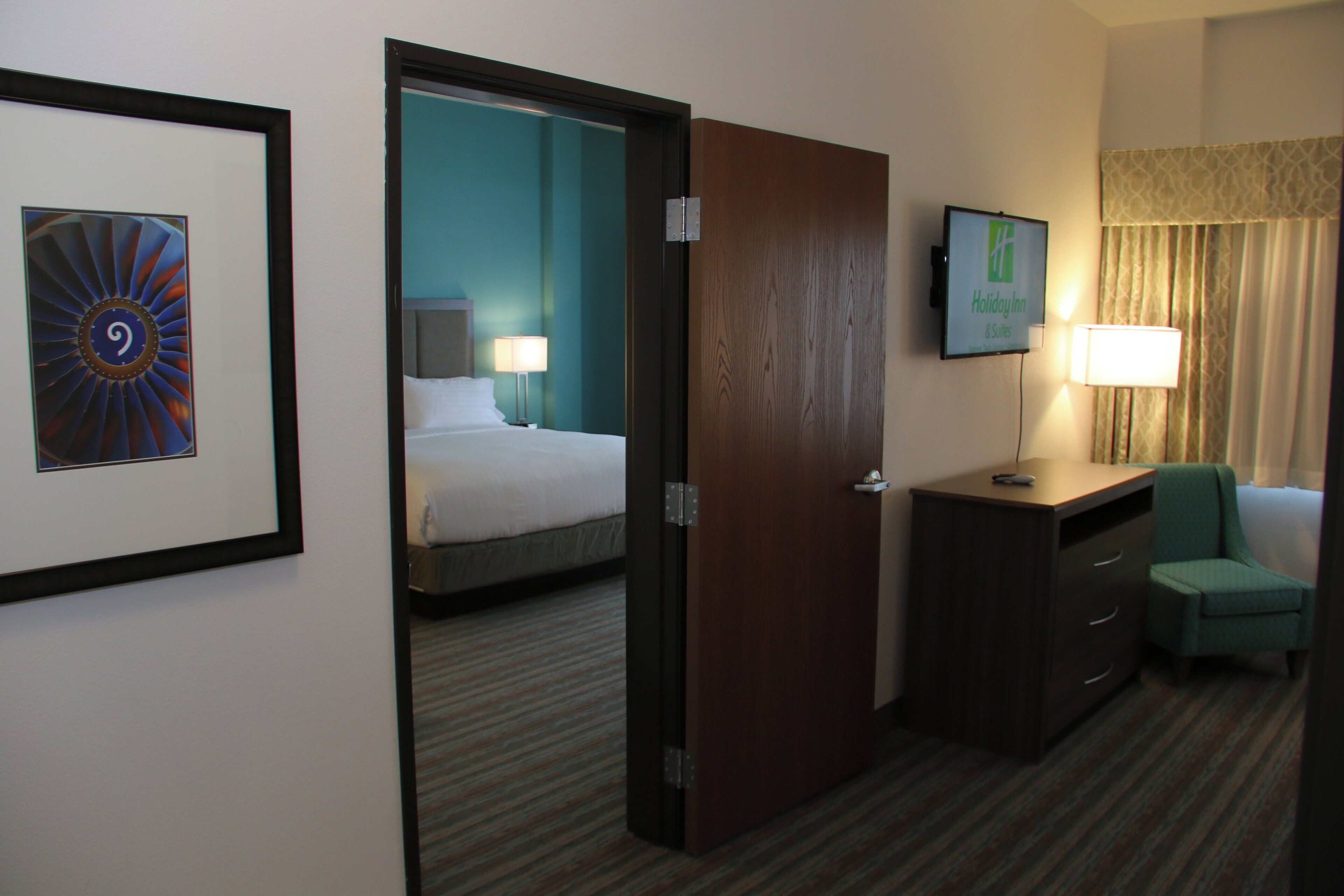 A hotel room with a bed, a desk, a chair, a TV, and a door leading to another room.