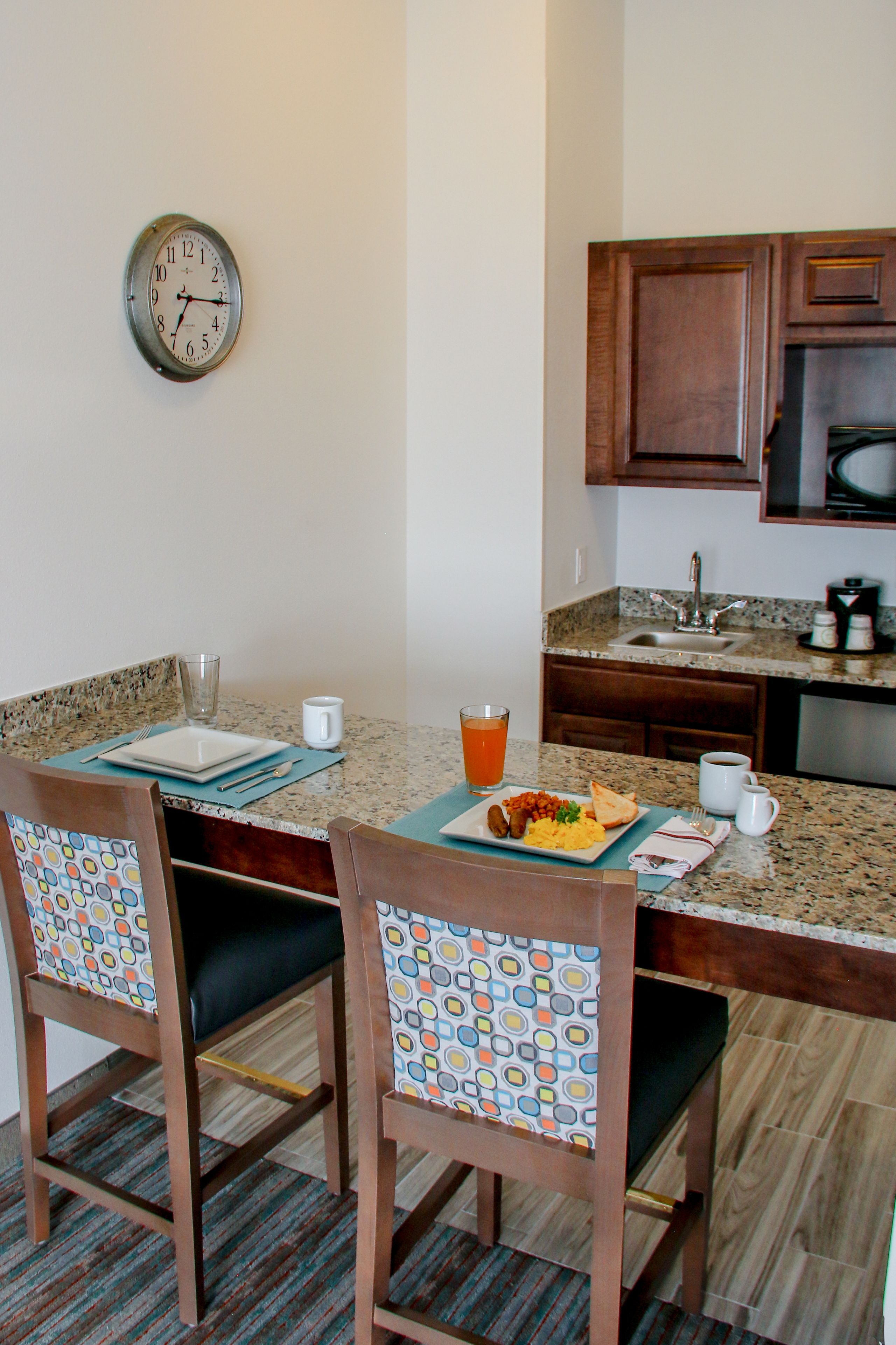 Un'area pranzo con un tavolo apparecchiato per due, sedie con rivestimenti decorati, una sveglia appesa al muro e una cucina sullo sfondo.