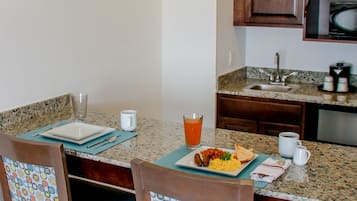 Un'area pranzo con un tavolo apparecchiato per due, sedie con rivestimenti decorati, una sveglia appesa al muro e una cucina sullo sfondo.