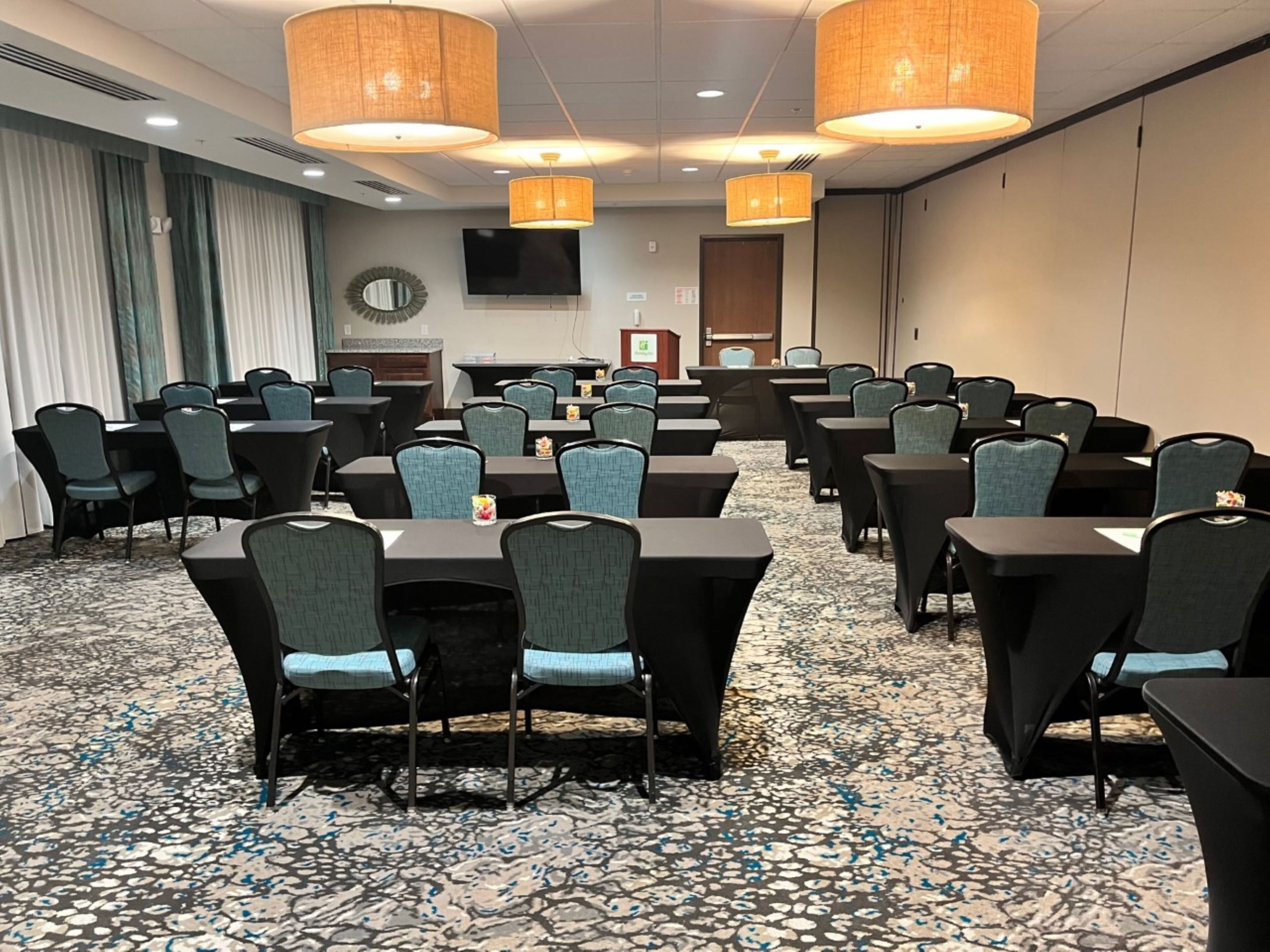 A conference room with multiple tables and chairs, a television on the wall, and a patterned carpet.