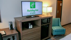 Une chambre d’hôtel équipée d’une télévision affichant le logo de Holiday Inn & Suites, d’un bureau avec une machine à café, d’une chaise et d’un lit.