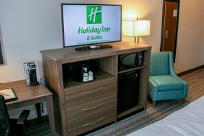 Une chambre d’hôtel équipée d’une télévision affichant le logo de Holiday Inn & Suites, d’un bureau avec une machine à café, d’une chaise et d’un lit.