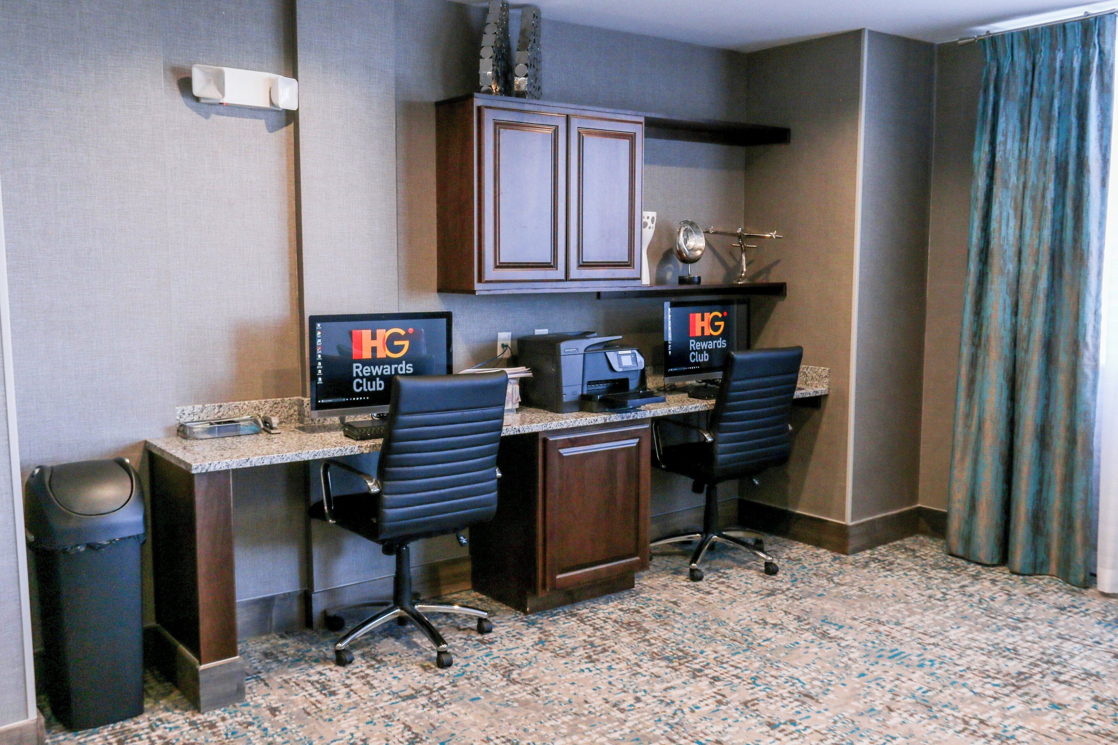 A hotel room with a desk, two chairs, a computer, a printer, and a trash can.