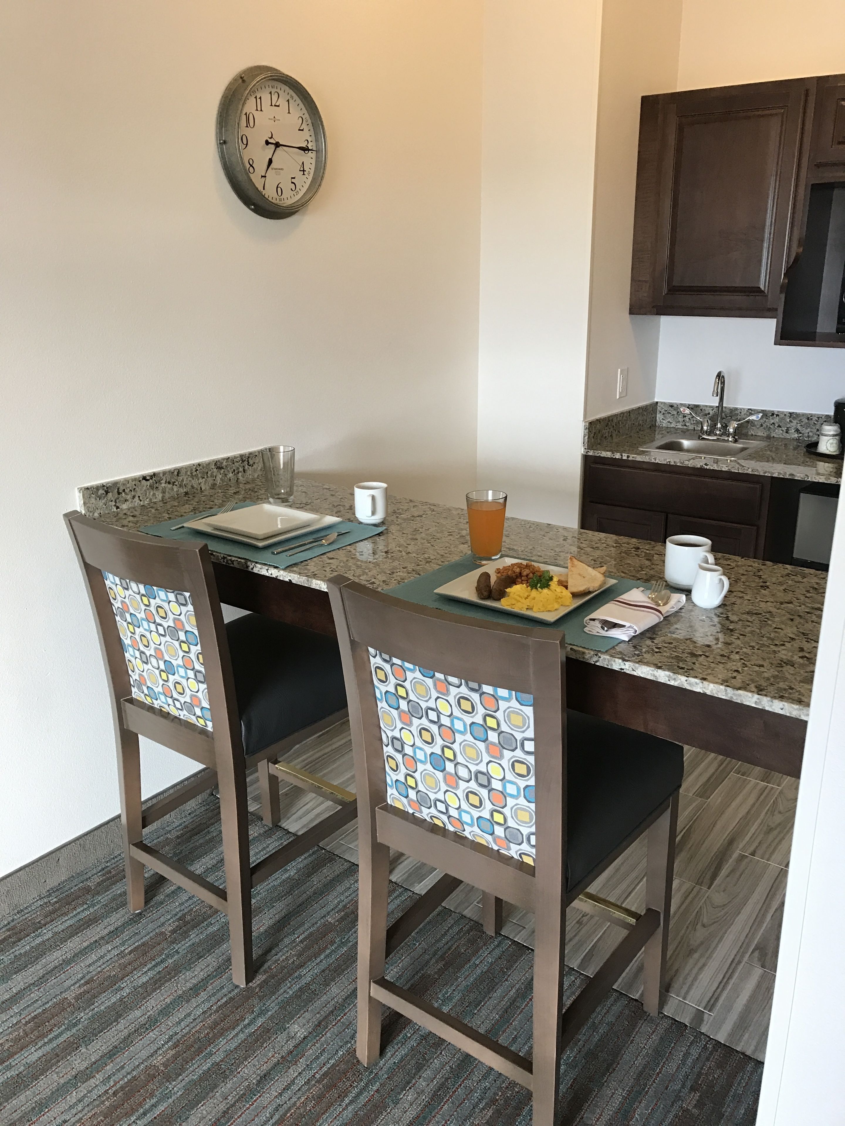 A dining area with a granite countertop, two chairs with patterned backs, a clock on the wall, and a kitchen in the background.