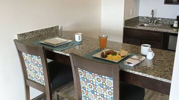 A dining area with a granite countertop, two chairs with patterned backs, a clock on the wall, and a kitchen in the background.