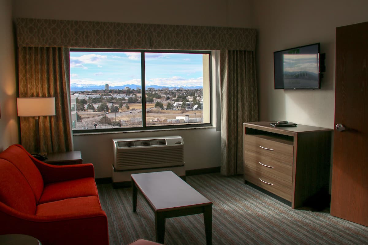 A hotel room with a large window, a red sofa, a wooden desk, and a television on the wall.