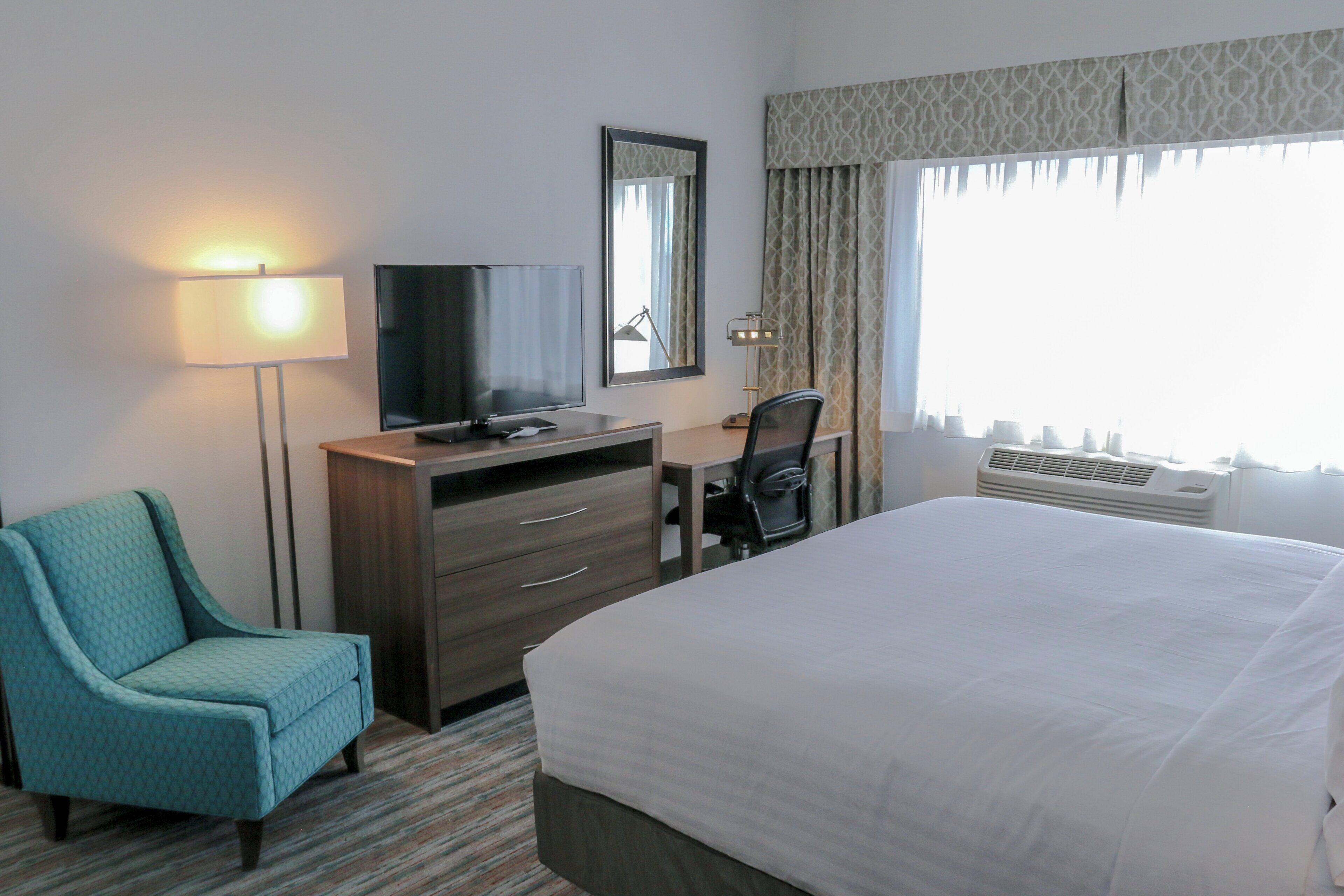A hotel room with a bed, a desk, a chair, a television, a lamp, and a large window with curtains.