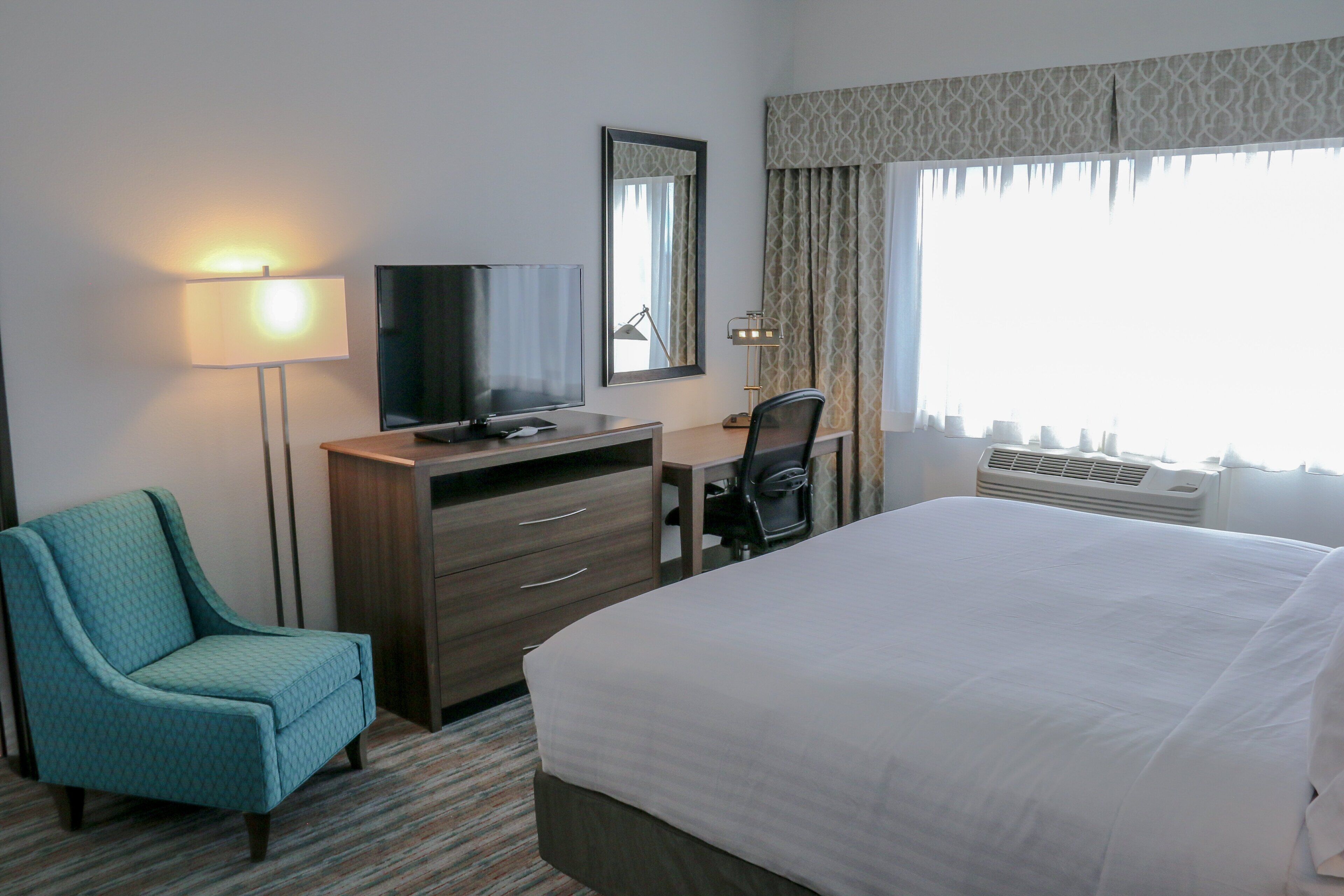 A hotel room with a bed, a desk with a television, a chair, a dresser, and a window with curtains.