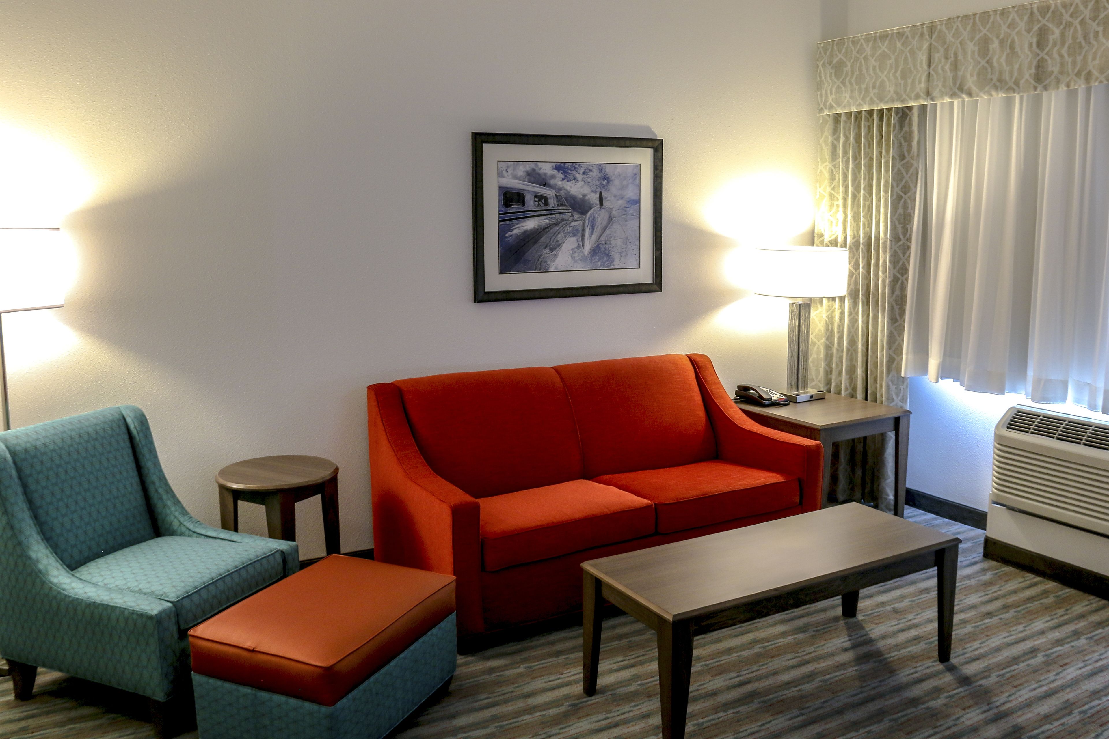 A hotel room with a red sofa, teal armchair, and a small table. There is a framed picture on the wall, a lamp, and a window with curtains.