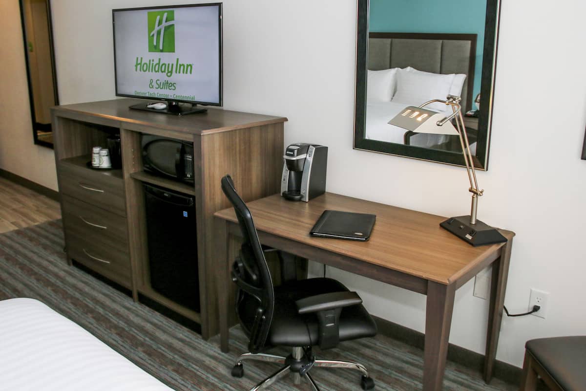 A hotel room with a wooden desk, a black office chair, a television displaying the Holiday Inn logo, a coffee maker, and a bedside lamp.