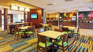 Een hotellobby met verschillende tafels en stoelen, een aan de muur bevestigde televisie en een balie met diverse spullen.