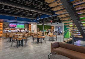 Un bar moderno con varias opciones de asientos, una pantalla mostrando deportes y una escalera con un muro de acento verde.